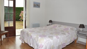 Une chambre à coucher avec un lit, une table de chevet, une lampe, une chaise et une vue sur un jardin, visible à travers une porte ouverte.