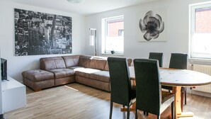 Een moderne woonkamer met een modulaire bank, een eettafel met stoelen, een groot stadslandschap schilderij en een bloemwerk op de muur.