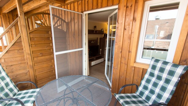 Una terraza de madera con una mesa redonda de metal y dos sillas a cuadros, una escalera de madera y un portón corredizo de vidrio que da a un espacio interior.