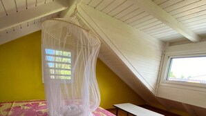 Ein Schlafzimmer mit einem Bett, das mit einer Bettdecke in Rosa und Gelb bezogen ist, einem weißen Baldachin, einem kleinen Tisch und einem Fenster mit Blick auf Grünflächen.