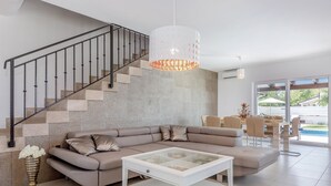 Un salon moderne avec un canapé d’angle, une table basse en verre, un escalier doté d’une rampe en métal et une grande fenêtre donnant sur l’extérieur.