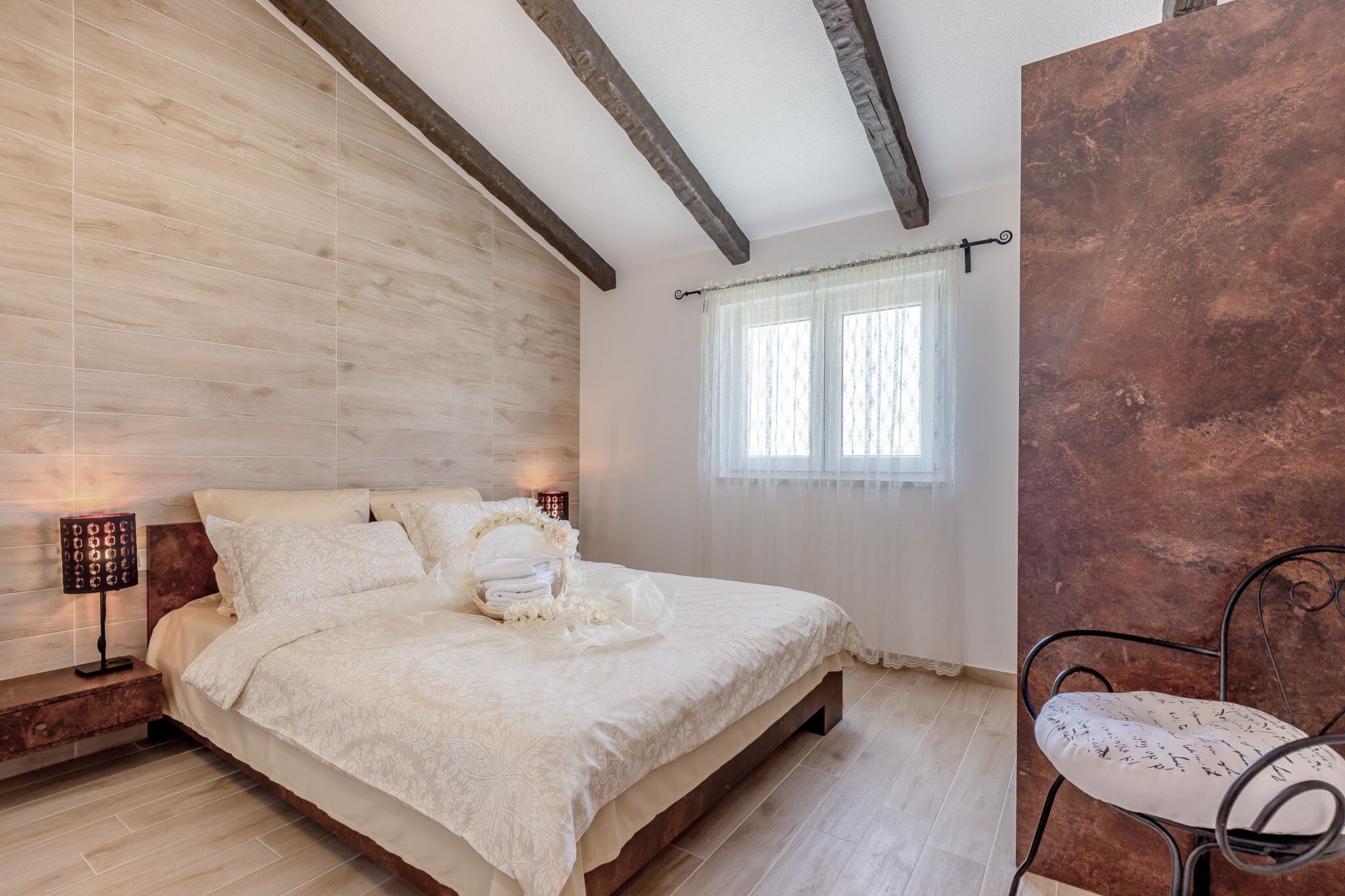A bedroom with a wooden headboard, a bed with a white bedspread, a bedside table with a lamp, and a window with sheer curtains.