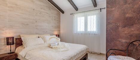 A bedroom with a wooden headboard, a bed with a white bedspread, a bedside table with a lamp, and a window with sheer curtains.