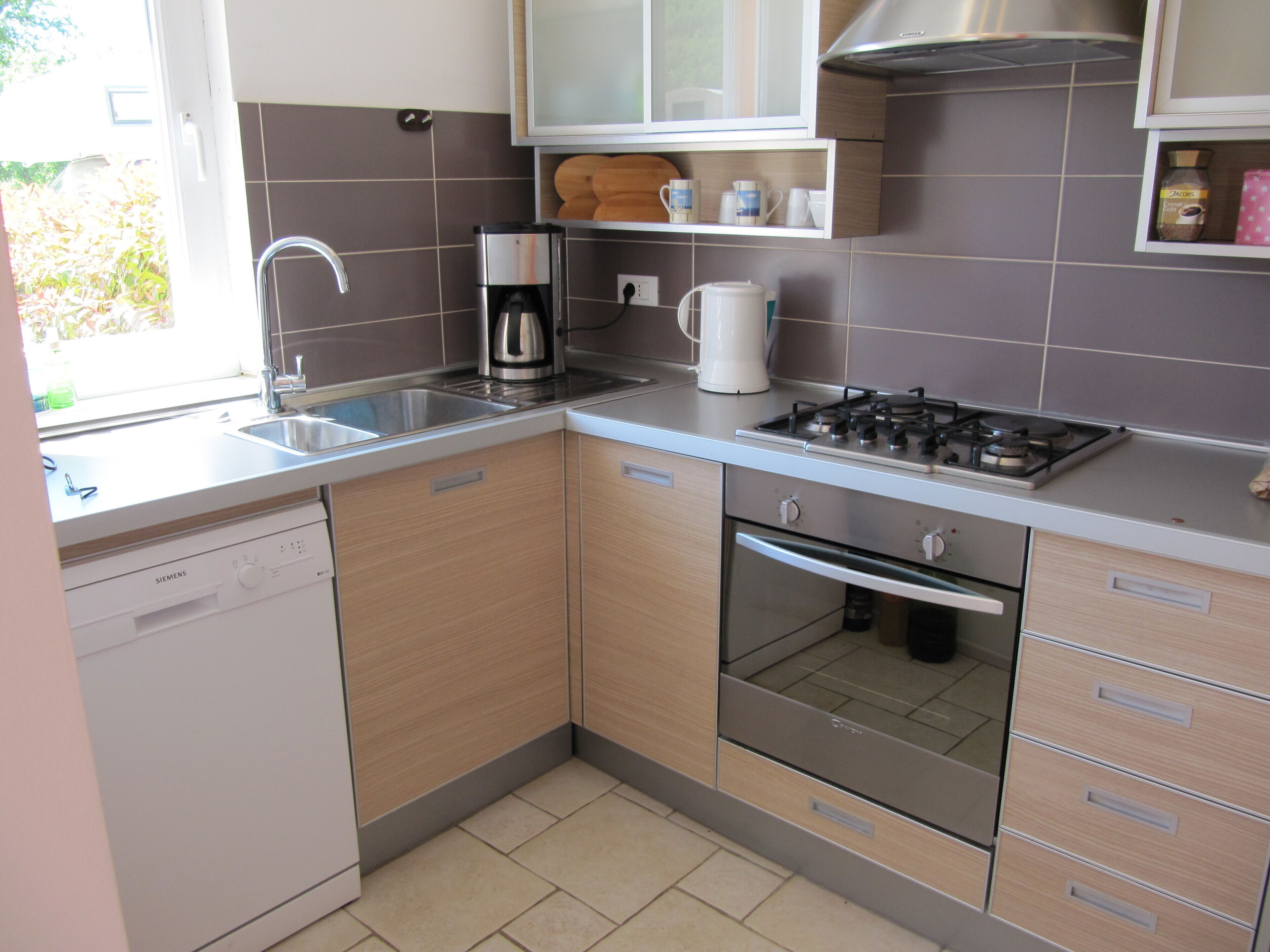 A modern kitchen with a dishwasher, sink, coffee maker, kettle, stove, oven, and wooden cabinets.