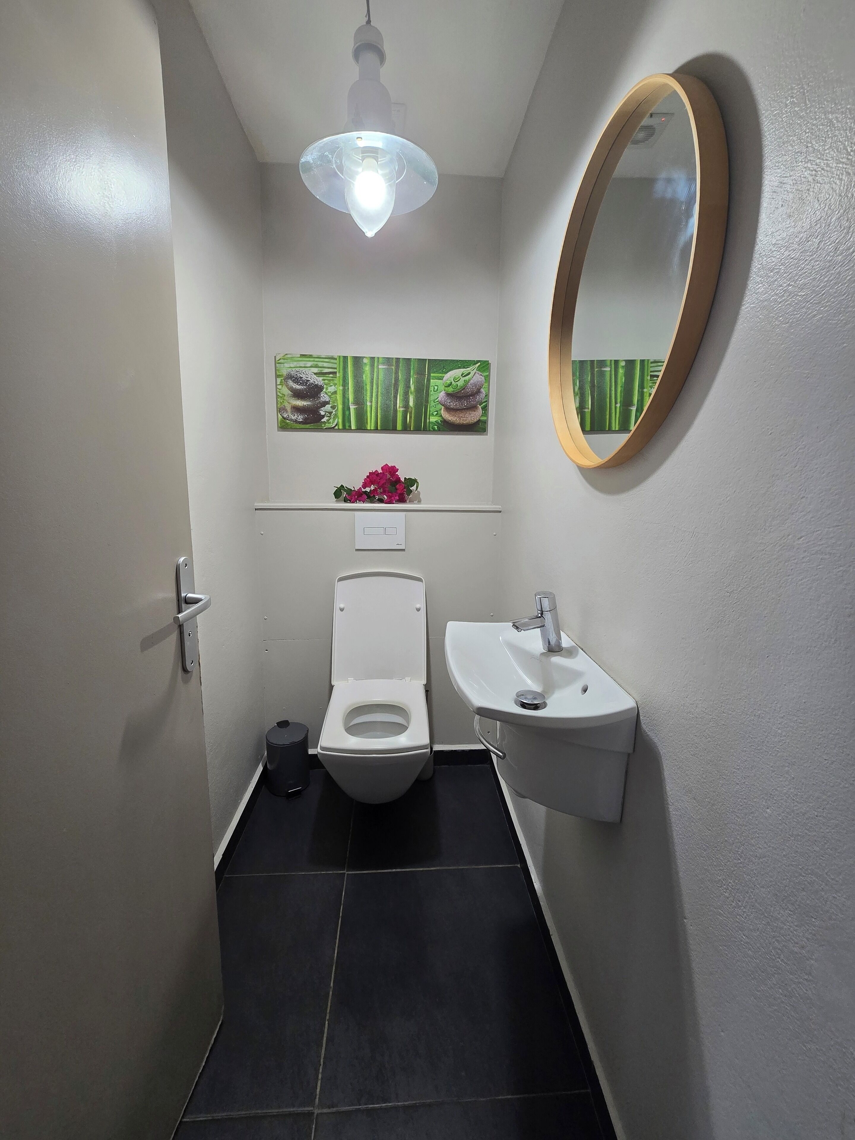 A small bathroom with a toilet, sink, and a mirror. There is a light fixture above the toilet and a shelf with a vase of flowers.