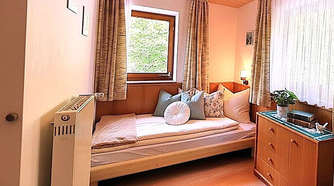 Una cama individual con almohadas, una mesita de noche, un aparador y una ventana con cortinas.
