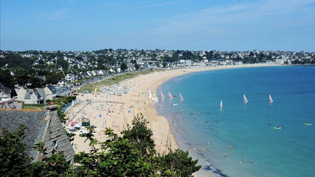 Une plage côtière avec des voiliers, des parasols et une vue dégagée sur le littoral.