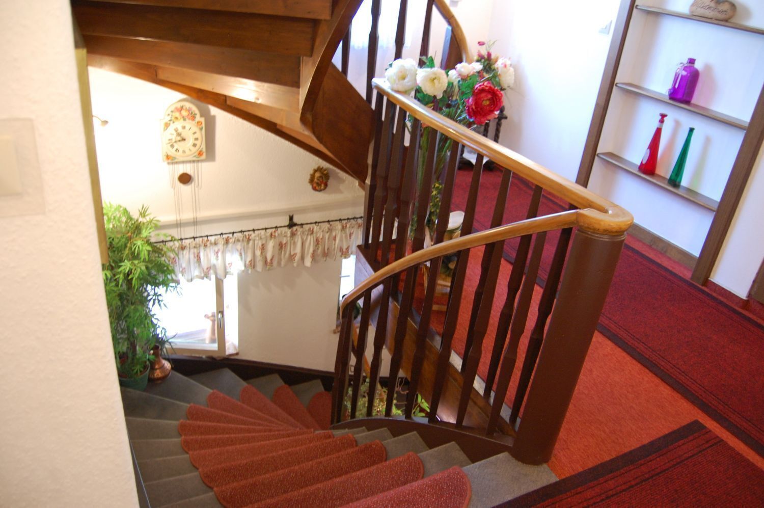 A wooden staircase with a handrail, a red carpeted landing, and a shelf with bottles.