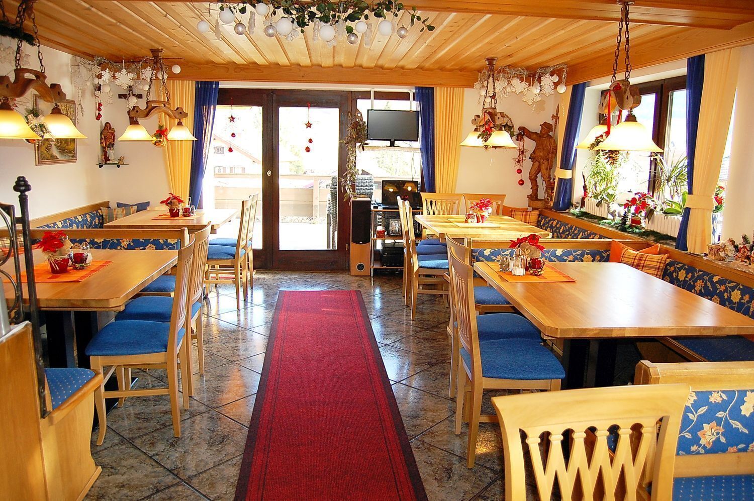 A cozy restaurant with wooden tables and blue upholstered benches, a red carpet leading to the entrance, and festive decorations.