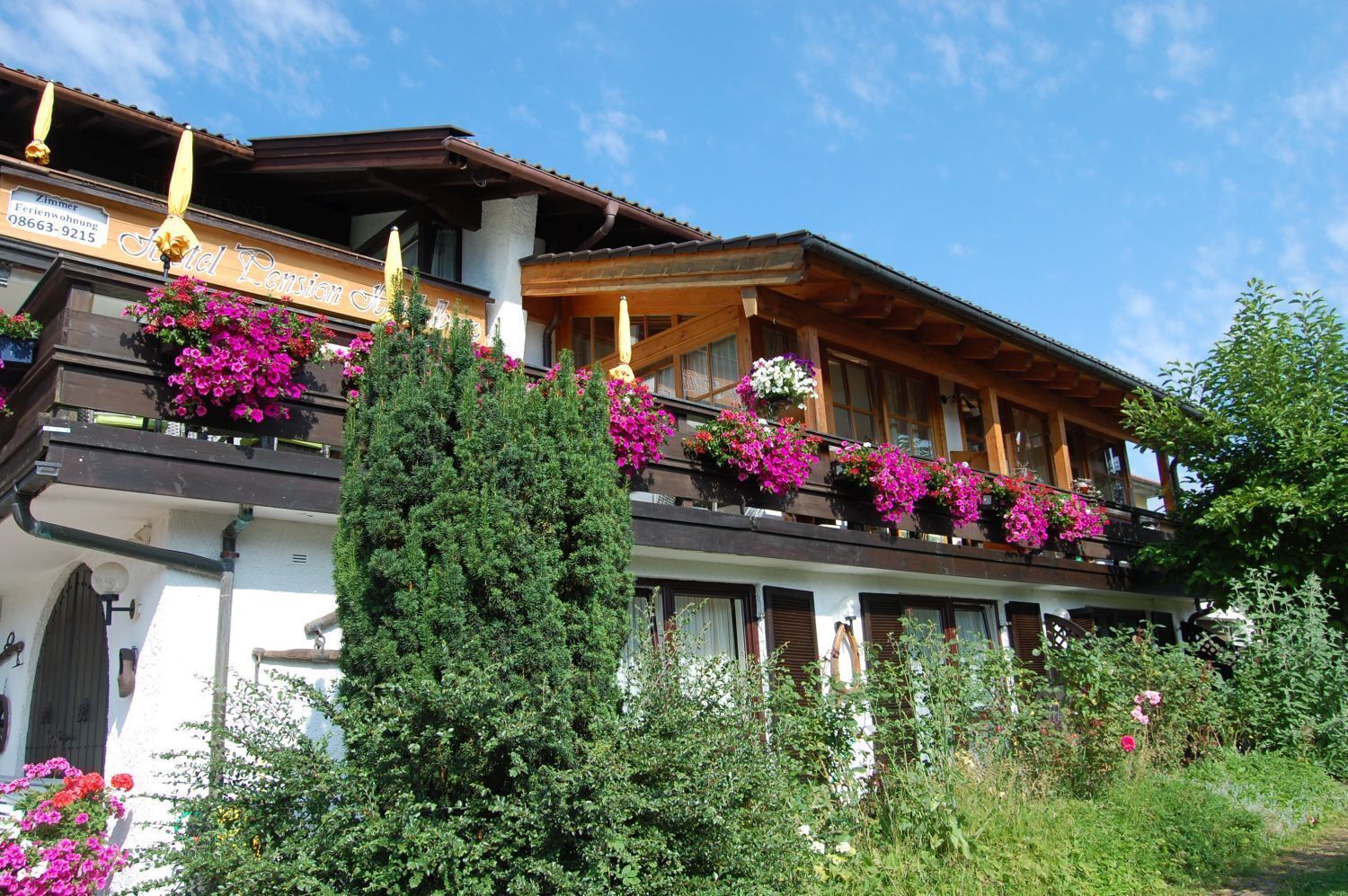 Un hôtel de style chalet traditionnel, doté de balcons ornés de fleurs aux couleurs vives, de poutres en bois et surplombant un ciel d’un bleu éclatant.