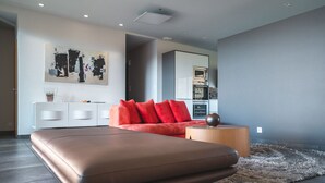 A modern bedroom with a large bed, a red sofa, a white console, and a kitchen area in the background.