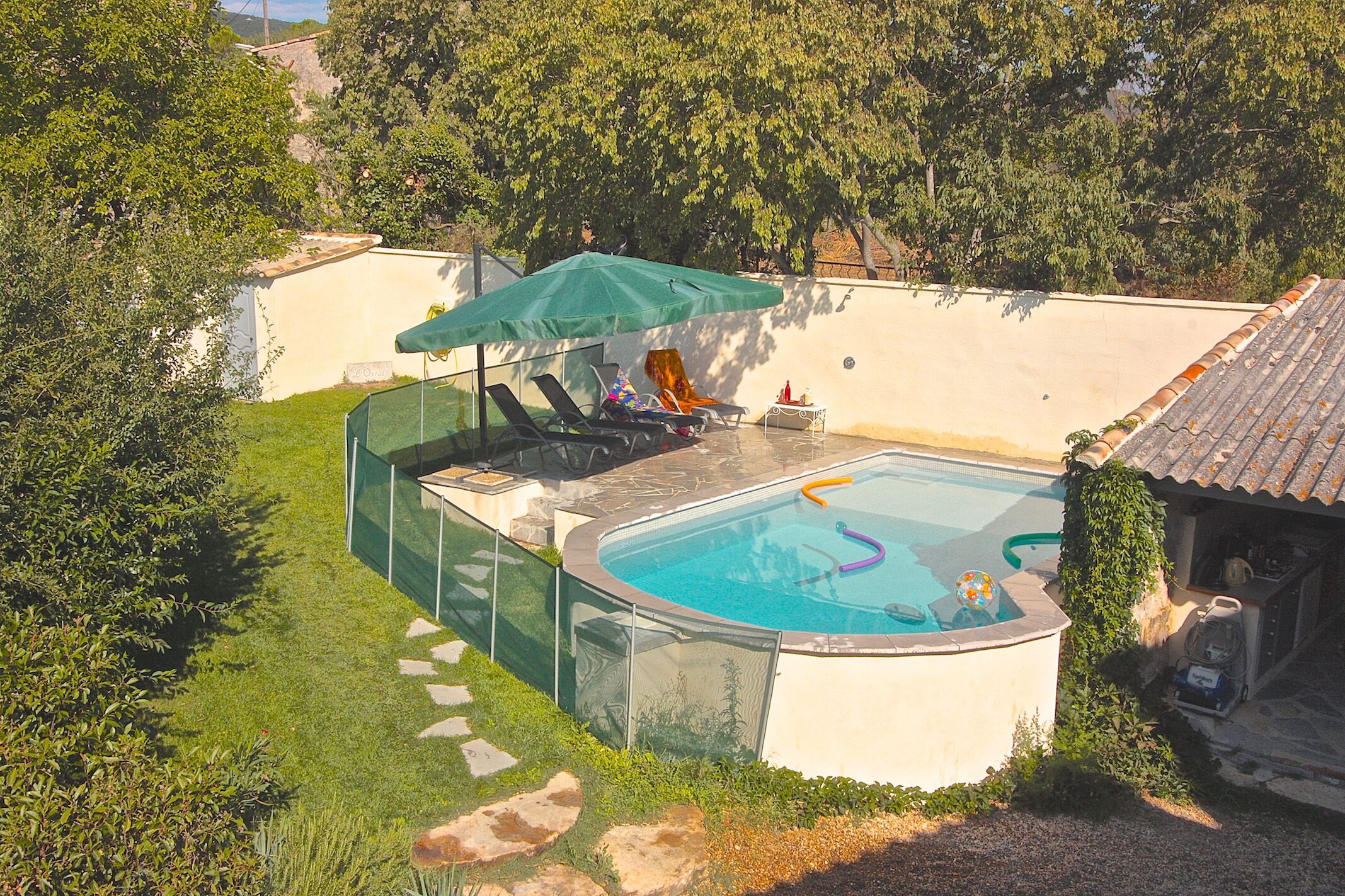 A swimming pool with a green umbrella, surrounded by a white wall and a grassy area with stepping stones.