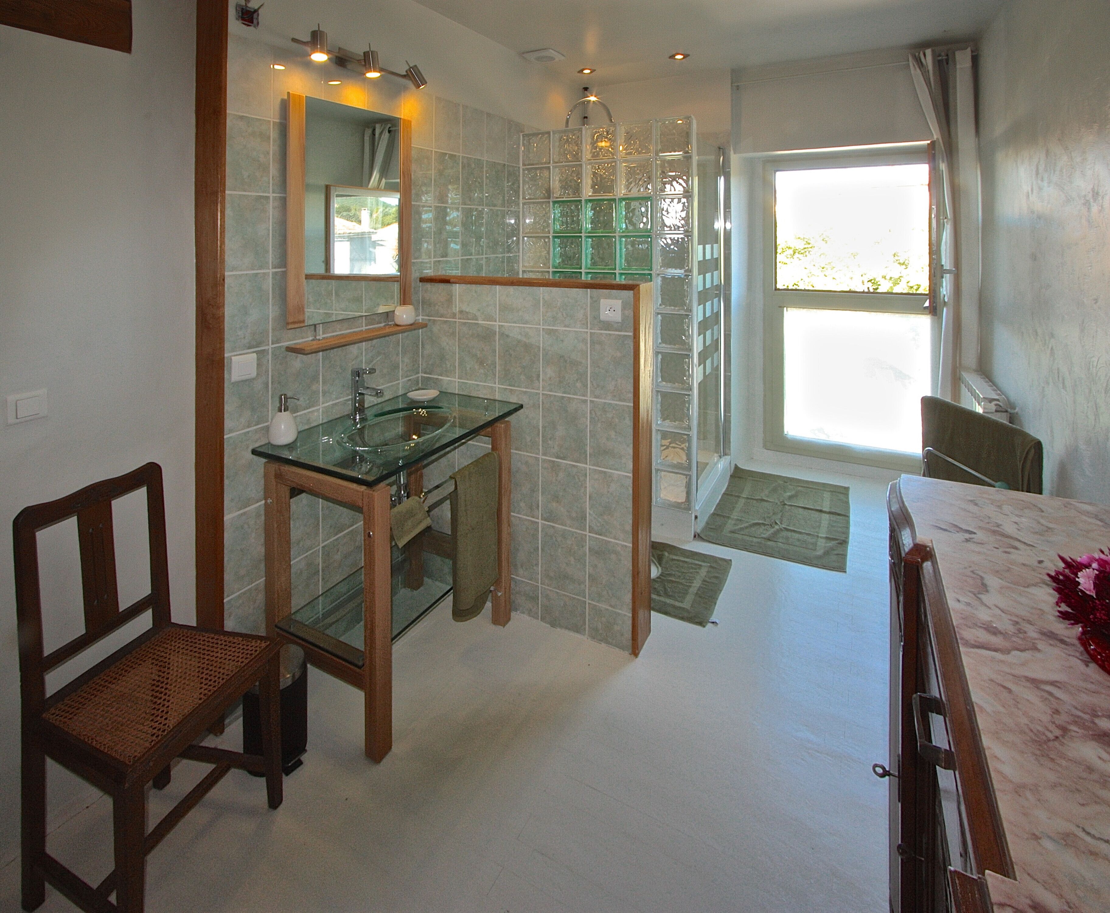 A bathroom with a glass sink, wooden chair, and a large window.