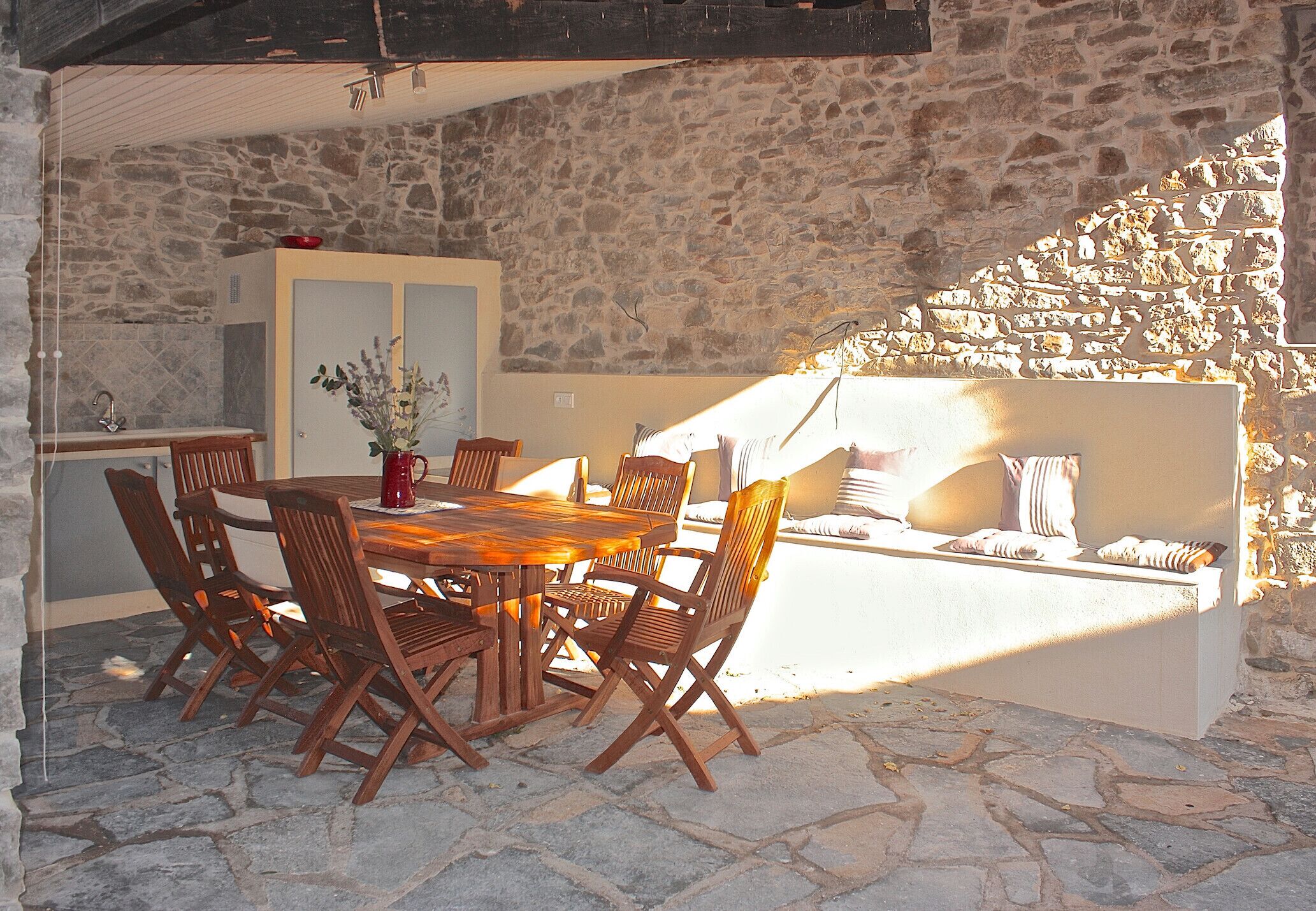 A stone-walled dining area with a wooden table and chairs, a white cabinet, and a stone bench with cushions.
