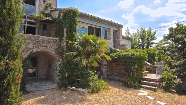 Un bâtiment en pierre doté d’un balcon, entouré de verdure et bordé d’un chemin de gravier.