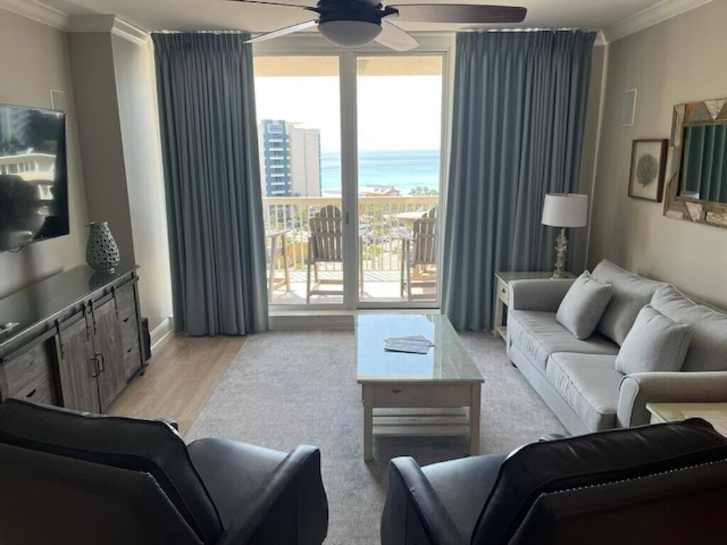 Una sala de estar con sofá, dos sillones, una mesa de centro, televisor y ventilador de techo. Hay un balcón con vista al océano y edificios.