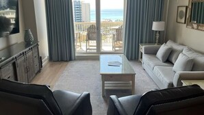 Una sala de estar con sofá, dos sillones, una mesa de centro, televisor y ventilador de techo. Hay un balcón con vista al océano y edificios.