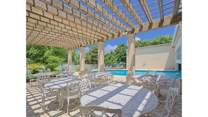 Relax by the pool under a shaded pergola, surrounded by lush greenery.