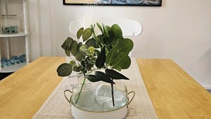 Una mesa con una planta en maceta dentro de un florero de vidrio, un mantel tejido y un cuadro abstracto enmarcado en la pared.