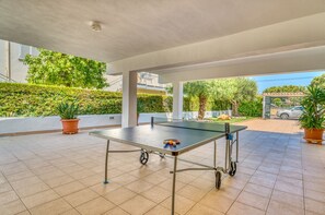 An outdoor ping pong table with wheels, surrounded by potted plants, under a covered area with tiled flooring.
