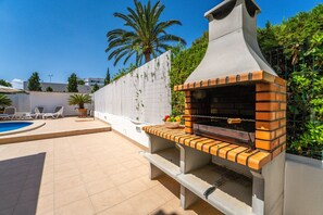 An outdoor brick oven grill with a chimney, surrounded by a pool area with lounge chairs and a palm tree.