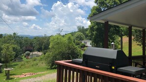 A covered outdoor deck with a grill, overlooking a lush green landscape and distant hills.