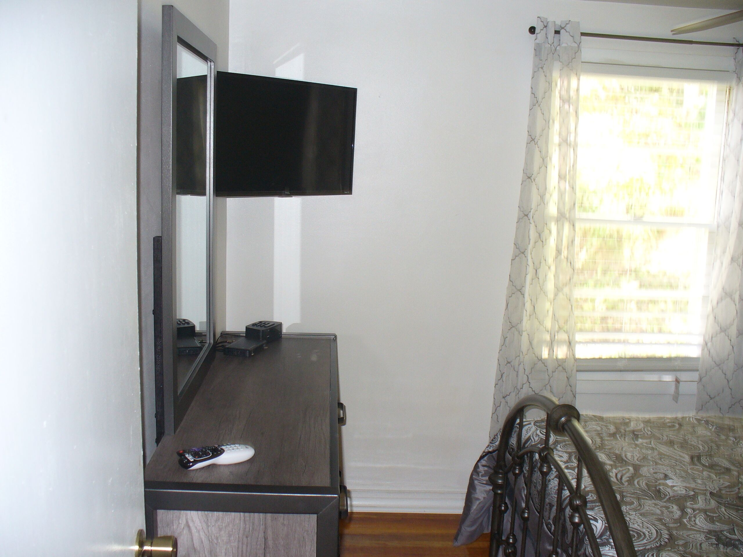 Quarto com penteadeira de madeira, espelho, televisão de tela plana fixada acima e cama com colcha estampada, além de janela com cortinas.