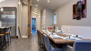 A modern dining area with a long wooden table set for six, a stainless steel refrigerator, and a view into a bright room.