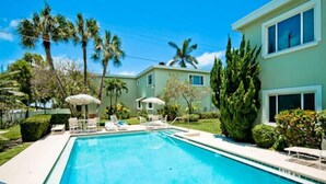 A swimming pool with lounge chairs and umbrellas, surrounded by palm trees and a building with large windows. - Gulf Beach Apartment - with Pool & Beach Access  (Holmes Beach)