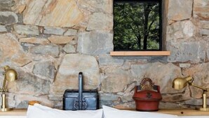 Una cama con ropa de cama blanca, pared de piedra, pequeña ventana con vista al bosque y dos lámparas de latón en una mesita de noche.