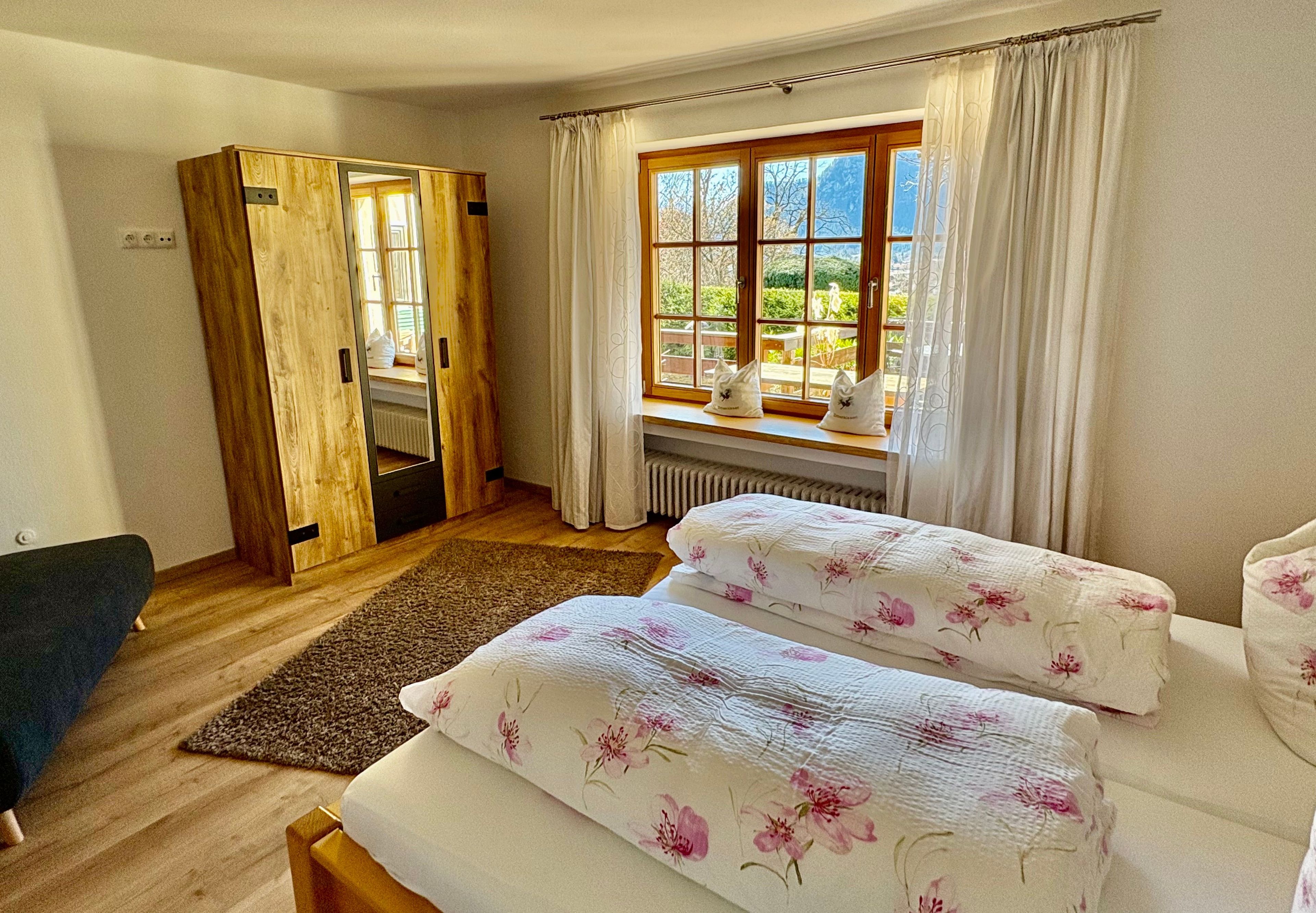 A bedroom with two beds, a wooden wardrobe, a window with curtains, and a view of greenery outside.