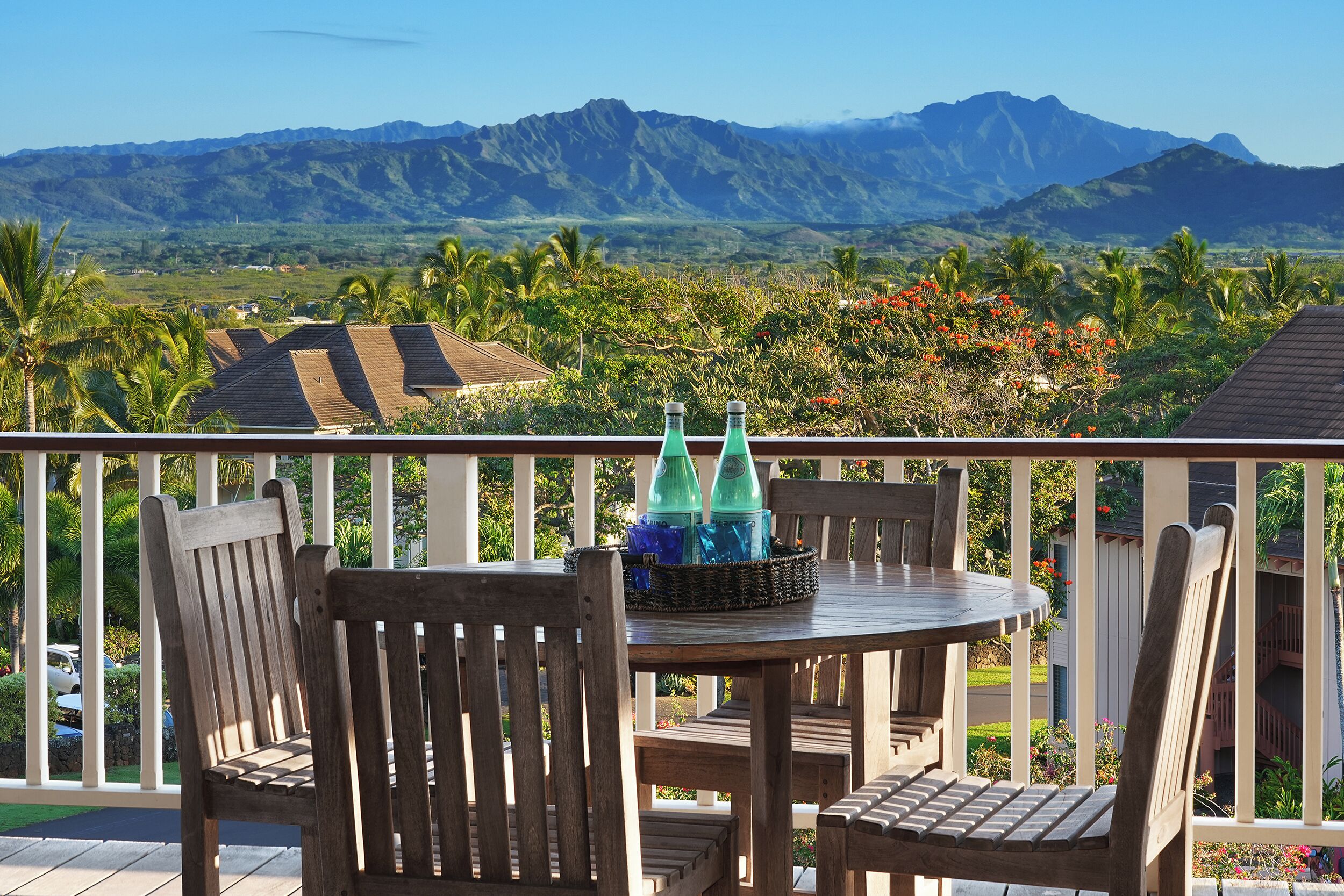 A wooden deck with a table and chairs, overlooking a mountainous landscape with lush greenery and red flowers.