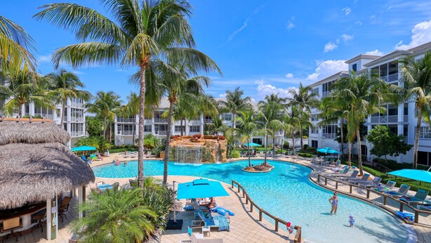 A resort with a swimming pool, lounge chairs, palm trees, and a thatched-roof structure. - Spacious Poolside End Unit! (Bonita Springs)