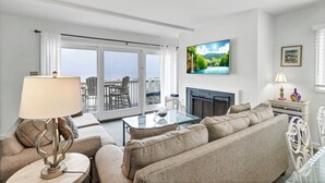 Een woonkamer met een bank, een glazen salontafel, een aan de muur bevestigde televisie en een open haard.