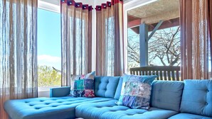 Un sofá seccional azul con cojines estampados, una mesa de centro de madera y cortinas de gasa con flecos.