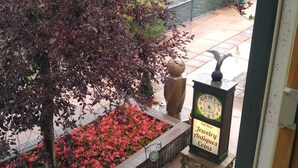 Blick aus einem Fenster auf eine Terrasse mit einer Uhr, einem Schild für Schmuck, Antiquitäten und Münzen sowie einer Bank.