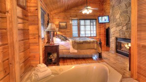 A log cabin bedroom with a stone fireplace, a large bed, a ceiling fan, and a television mounted on the wall.