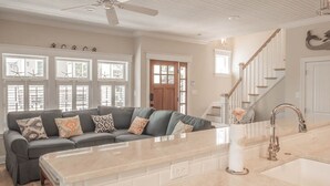 A spacious living room with a large island, a sofa with patterned cushions, and a view of the kitchen through a glass door. - CORNER BEACH HOUSE - COTTAGE (Cape May)