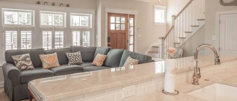 A spacious living room with a large island, a sofa with patterned cushions, and a view of the kitchen through a glass door.