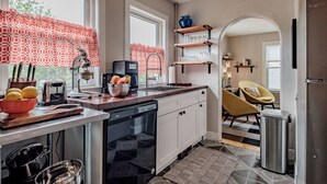 Een moderne keuken met witte kasten, een zwarte vaatwasser en een rood gordijn voor het raam. Boven de gootsteen bevinden zich houten planken en er staat een stoel in het aangrenzende eetgedeelte.