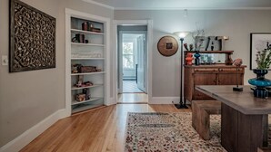 Een kamer met houten vloer, een boekenkast, een eettafel met banken, een lamp en een decoratief wandobject.