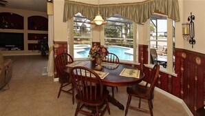 Un coin repas avec une table ronde, des chaises et un vase de fleurs. On peut apercevoir une piscine à travers les fenêtres.