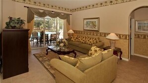 Un salon avec un canapé, une table basse et des lampes. On aperçoit un coin repas avec des chaises et une table à travers une porte vitrée.