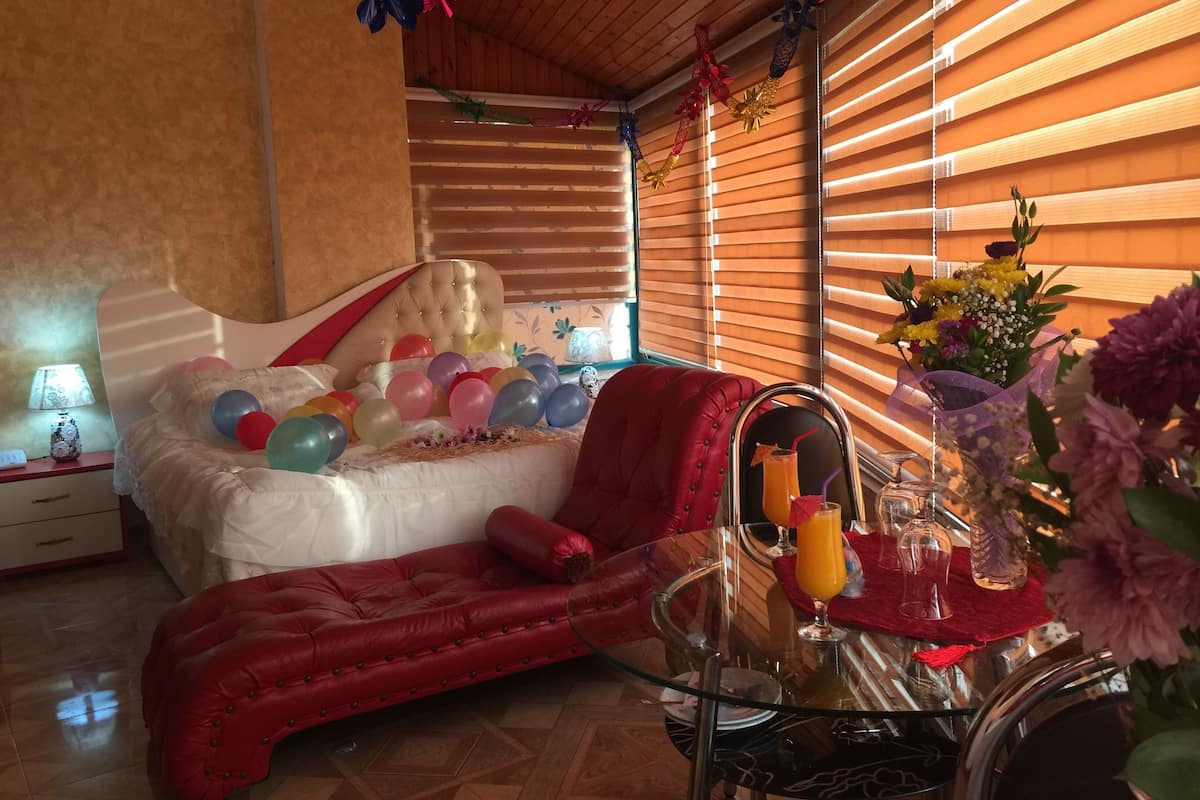 Um quarto com uma cama, uma poltrona vermelha, uma mesa de vidro com bebidas e uma janela com persianas.