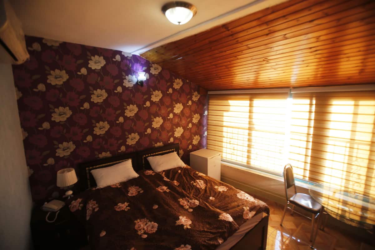 Quarto com papel de parede floral, teto de madeira, cama com colcha estampada, mesa de cabeceira com abajur e cadeira próxima a janela com persianas.
