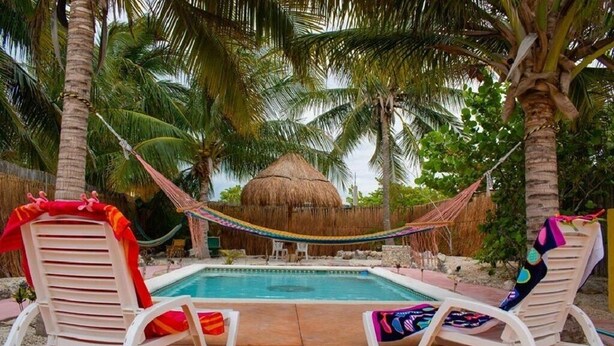 A pool area with two white lounge chairs, a hammock, and a thatched-roof structure.