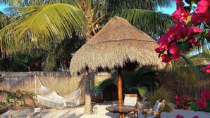A thatched-roof outdoor seating area with a table and chairs, surrounded by palm trees and tropical plants.