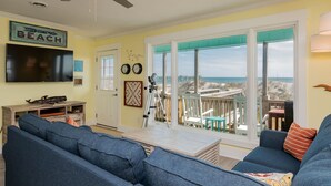 Un soggiorno con un'ampia televisione, divani blu, un tavolino in legno e un balcone con vista sulla spiaggia.