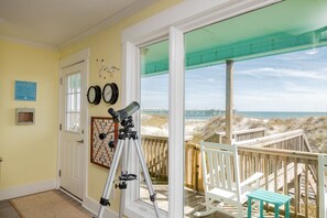 A room with a telescope, a framed board game, and a door leading to a balcony with a view of the beach and pier.