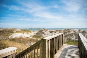 A wooden boardwalk leading to a beach with dunes and a pier in the background.